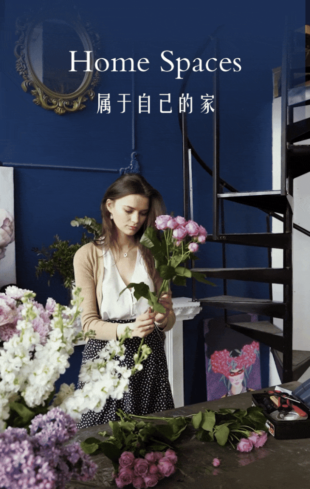 A woman arranges pink roses and white flowers in a room with a spiral staircase and blue walls.