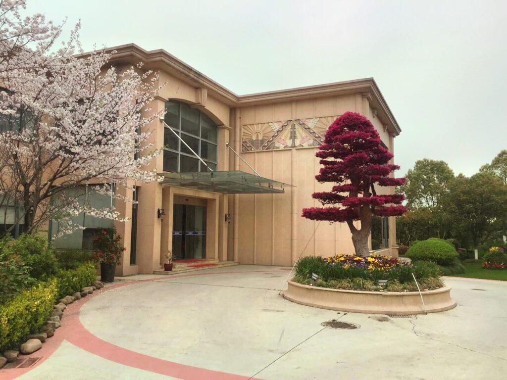 A beige building with a large glass entrance is flanked by a blooming cherry tree and a red-leafed ornamental tree.