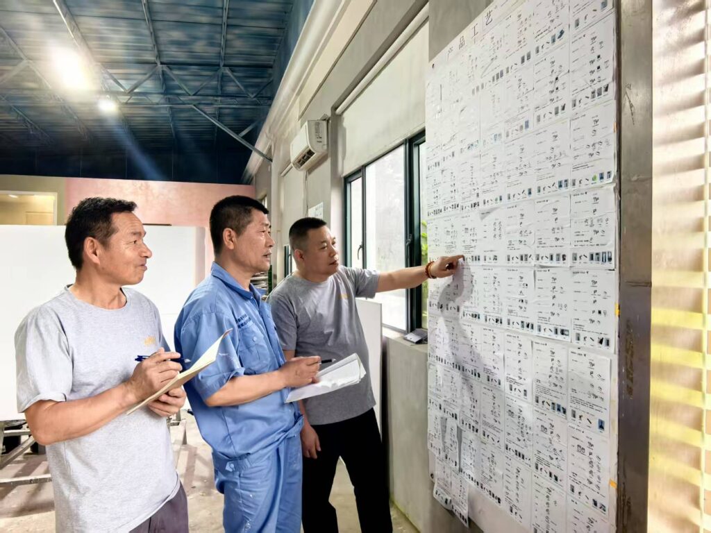 Three men review a large wall chart with many small images and text in a workshop setting.