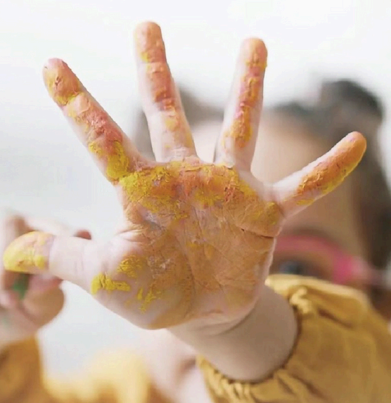 A child's hand is covered in yellow and orange paint, held up against a bright background.