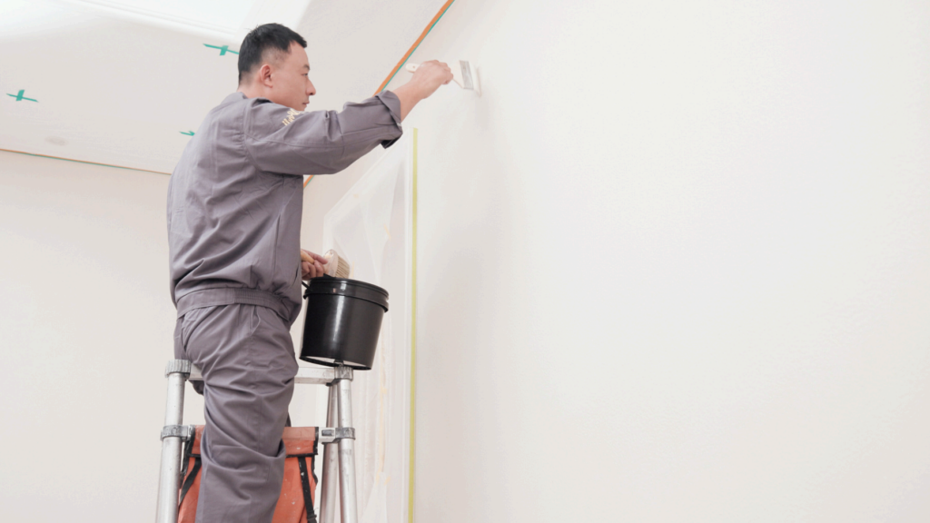A painter in a grey uniform stands on a ladder, applying white paint to a wall with a brush.
