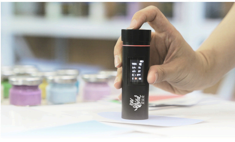 A hand holds a black device with a digital screen displaying color data, with colorful jars in the background.