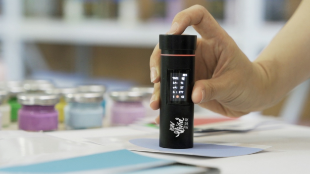 A hand holds a black digital colorimeter with a screen displaying color data, near small jars of colorful pigments.