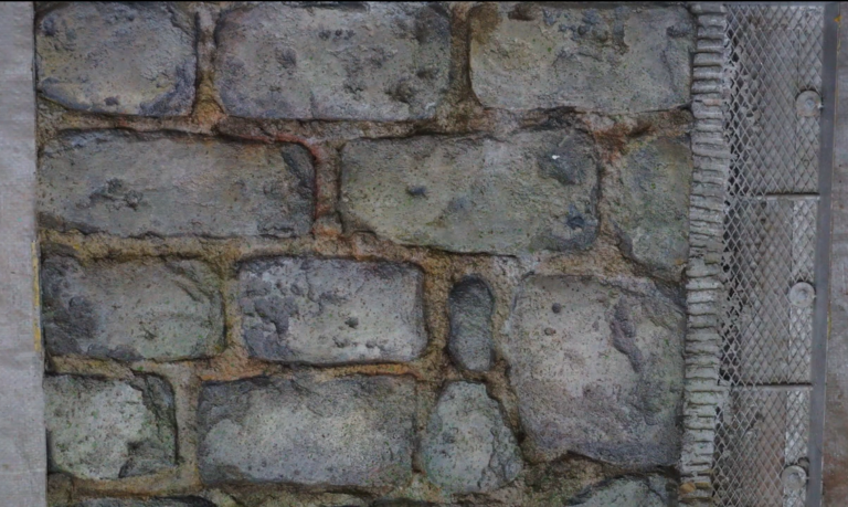 Roughly textured, uneven stones are mortared together, with a metal mesh on the right edge.