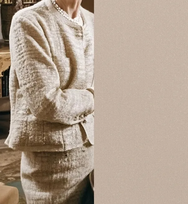 A person wears a textured beige tweed suit and pearl necklace, posing with arms crossed.