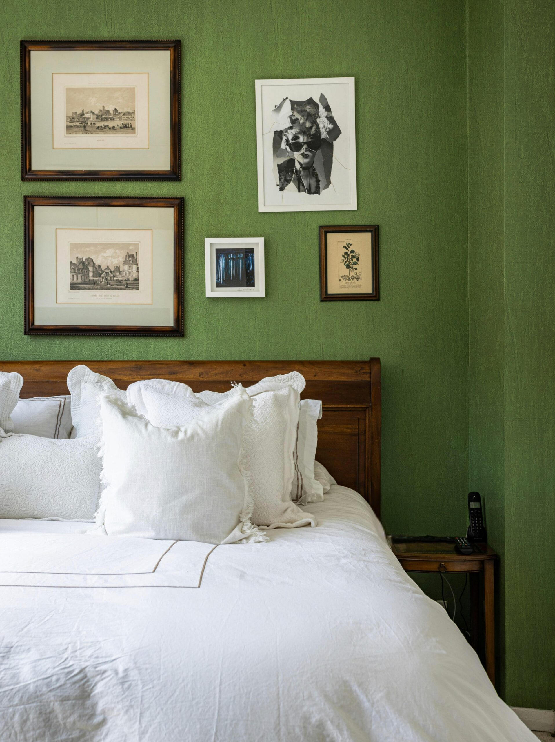 A white duvet covers a bed with a wooden headboard against a textured green wall adorned with antique prints.