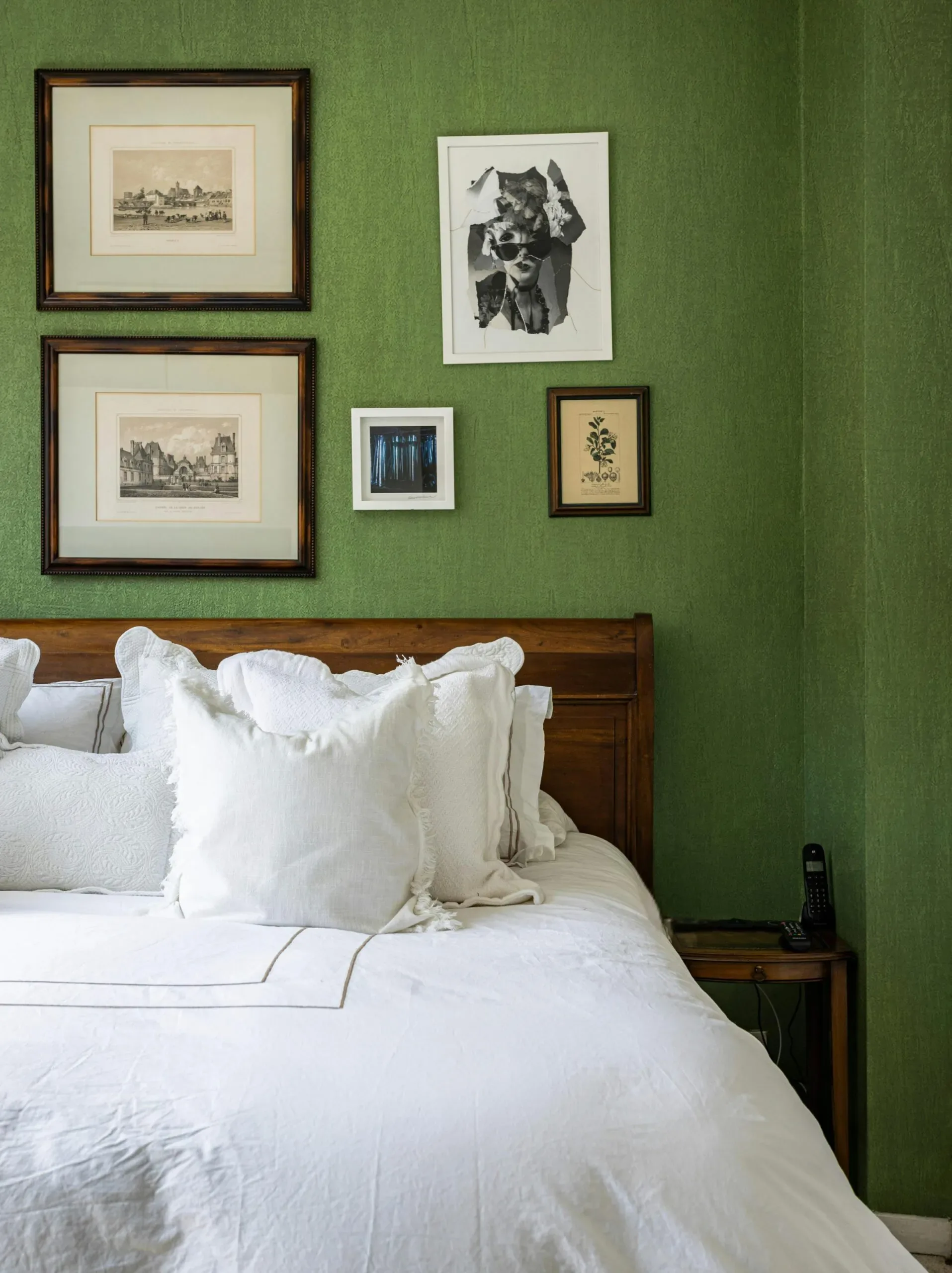 A white duvet covers a bed with a wooden headboard against a textured green wall adorned with antique prints.