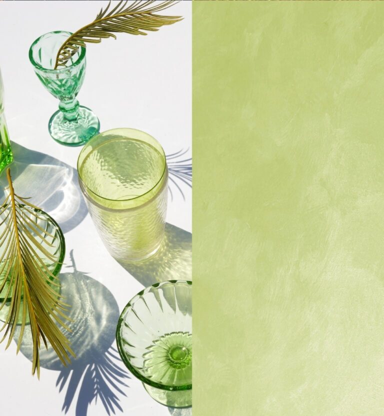 Green glassware and palm fronds cast shadows on a white surface next to a textured green background.