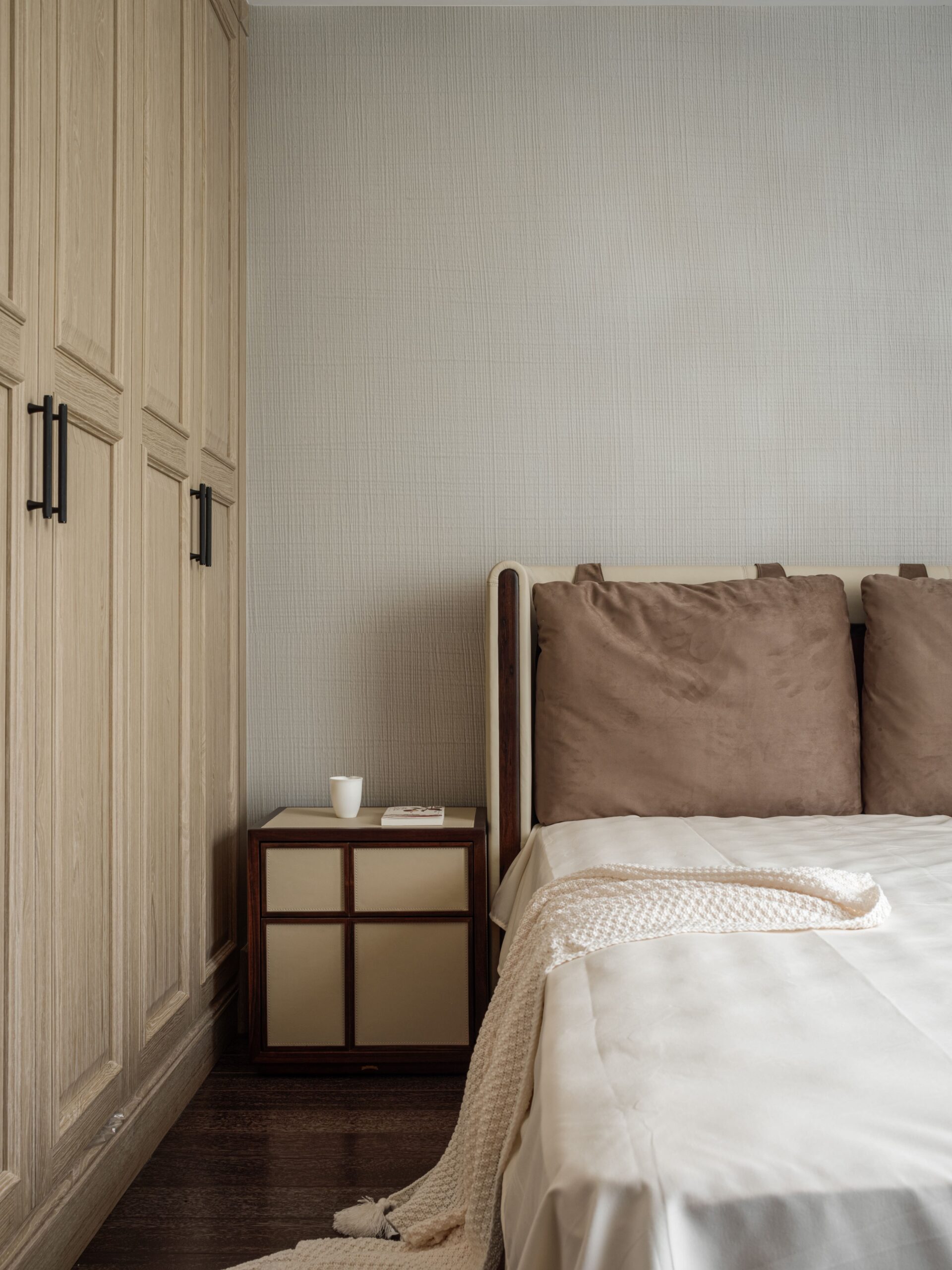 A minimalist bedroom features a wooden wardrobe, a bedside table with a cup, and a bed with brown pillows and a white throw.