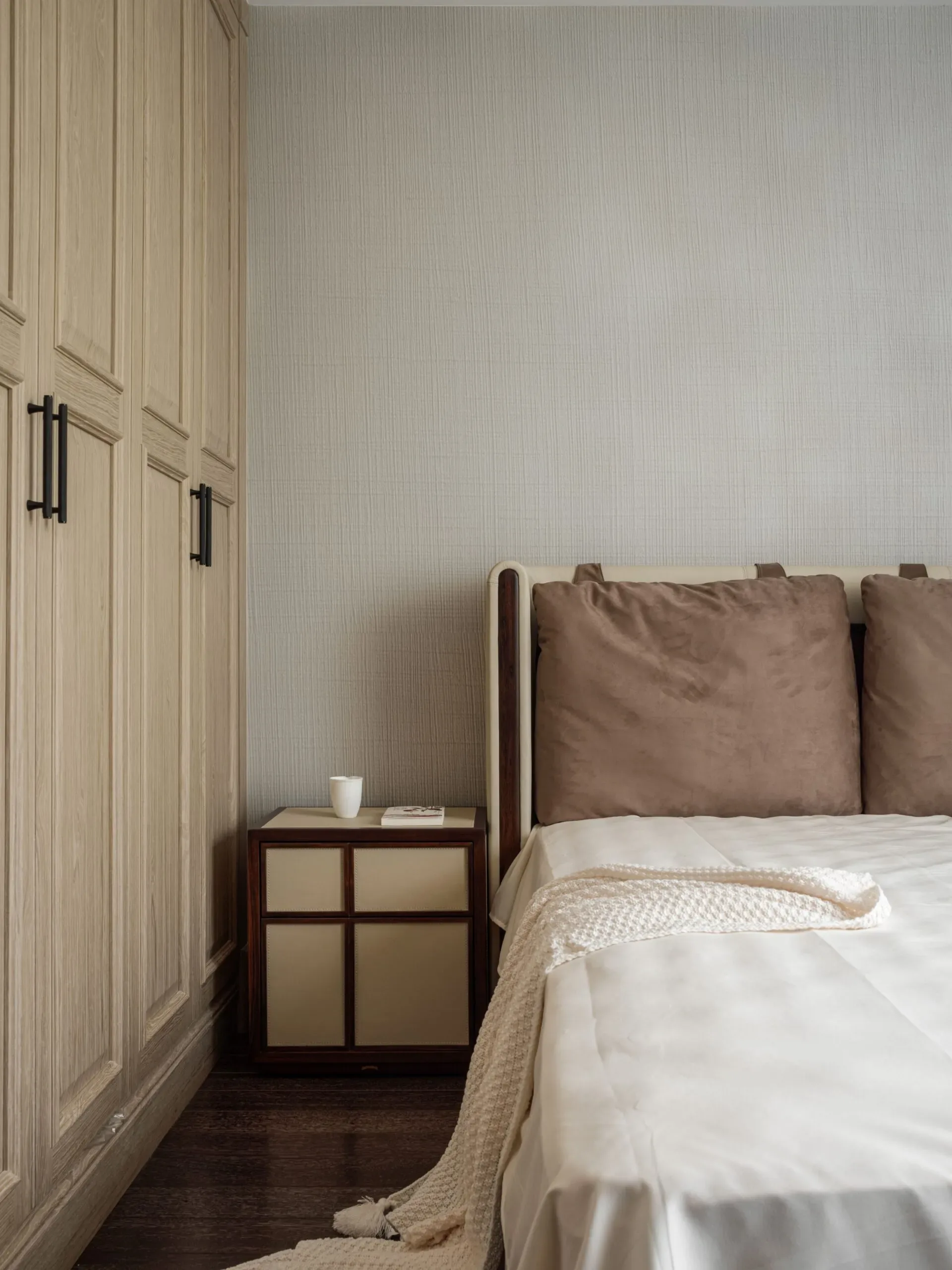 A minimalist bedroom features a wooden wardrobe, a bedside table with a cup, and a bed with brown pillows and a white throw.