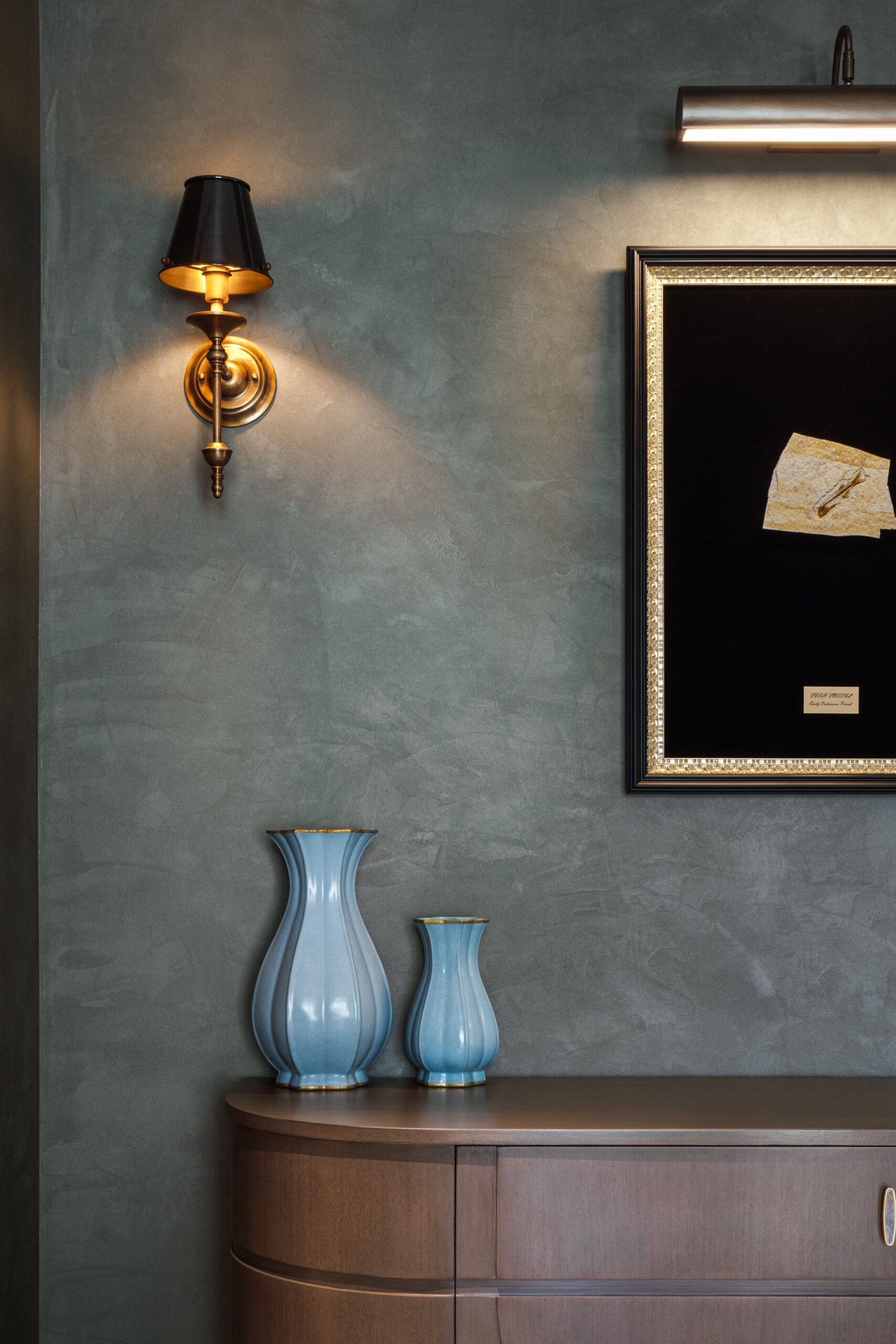 Two blue vases sit on a wooden dresser beneath a framed artwork and a wall sconce.