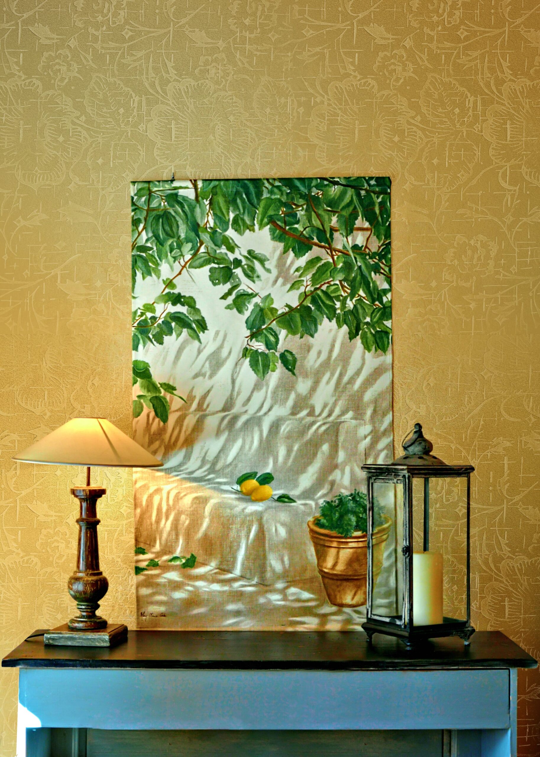 A still life painting of lemons and greenery hangs above a table with a lamp and lantern.